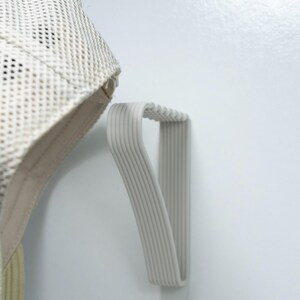 May include: A light gray, ribbed wall-mounted hat rack. The rack is made of a flexible material and is designed to hold a hat. A beige mesh hat is partially visible, hanging from the rack.