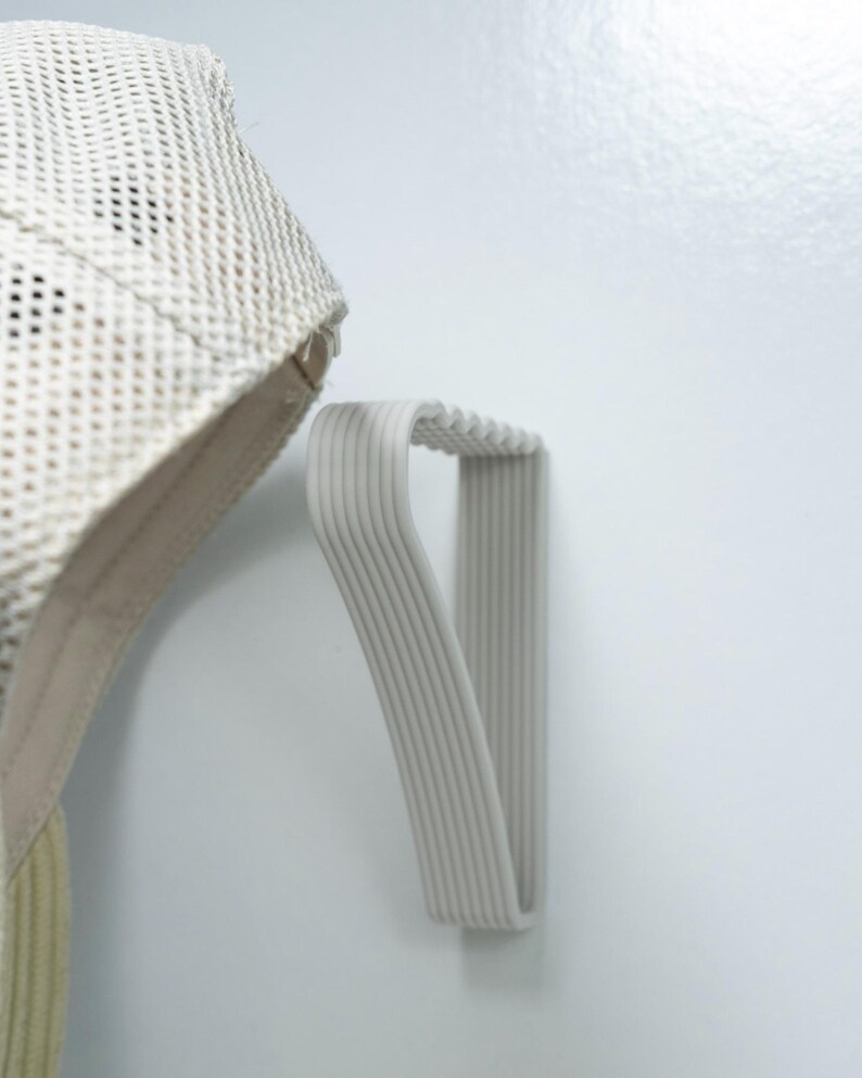 May include: A light gray, ribbed wall-mounted hat rack. The rack is made of a flexible material and is designed to hold a hat. A beige mesh hat is partially visible, hanging from the rack.