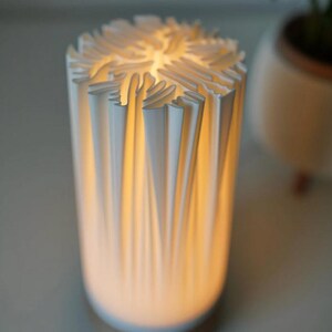 Puede incluir: Una l&aacute;mpara cil&iacute;ndrica blanca con un dise&ntilde;o &uacute;nico. El exterior de la l&aacute;mpara presenta una serie de elementos verticales plegados que crean una superficie texturizada. El interior brilla con una luz c&aacute;lida e invitadora.