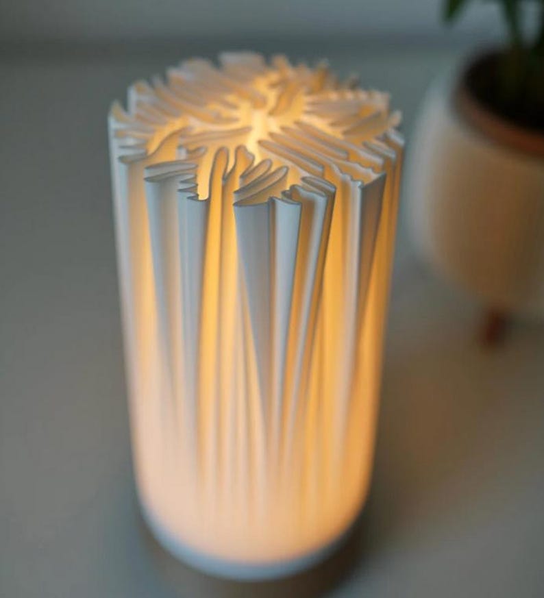 Puede incluir: Una l&aacute;mpara cil&iacute;ndrica blanca con un dise&ntilde;o &uacute;nico. El exterior de la l&aacute;mpara presenta una serie de elementos verticales plegados que crean una superficie texturizada. El interior brilla con una luz c&aacute;lida e invitadora.