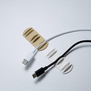 May include: Two silicone cable organisers, one beige and one white, each featuring three parallel raised ridges to secure cords. A white USB-C charging cable is threaded through the beige organiser, and a black USB-C cable is held by the white one.