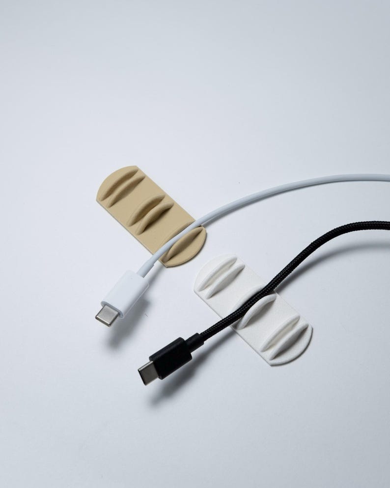 May include: Two silicone cable organisers, one beige and one white, each featuring three parallel raised ridges to secure cords. A white USB-C charging cable is threaded through the beige organiser, and a black USB-C cable is held by the white one.