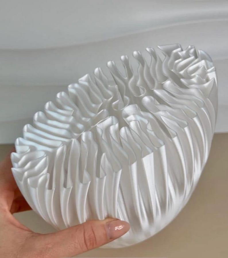 May include: A white, semi-transparent, decorative bowl with a unique, pleated design. The bowl has a rounded shape and is held by a hand. The pleats create a wave-like pattern, adding texture and visual interest to the piece.