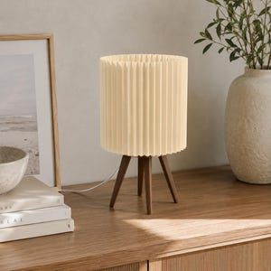 Puede incluir: Una l&aacute;mpara de mesa con una pantalla cil&iacute;ndrica plisada de color crema, sostenida por tres patas de madera de color marr&oacute;n oscuro. La l&aacute;mpara incluye un cable de alimentaci&oacute;n blanco que sale de la base, con un dise&ntilde;o estructurado.