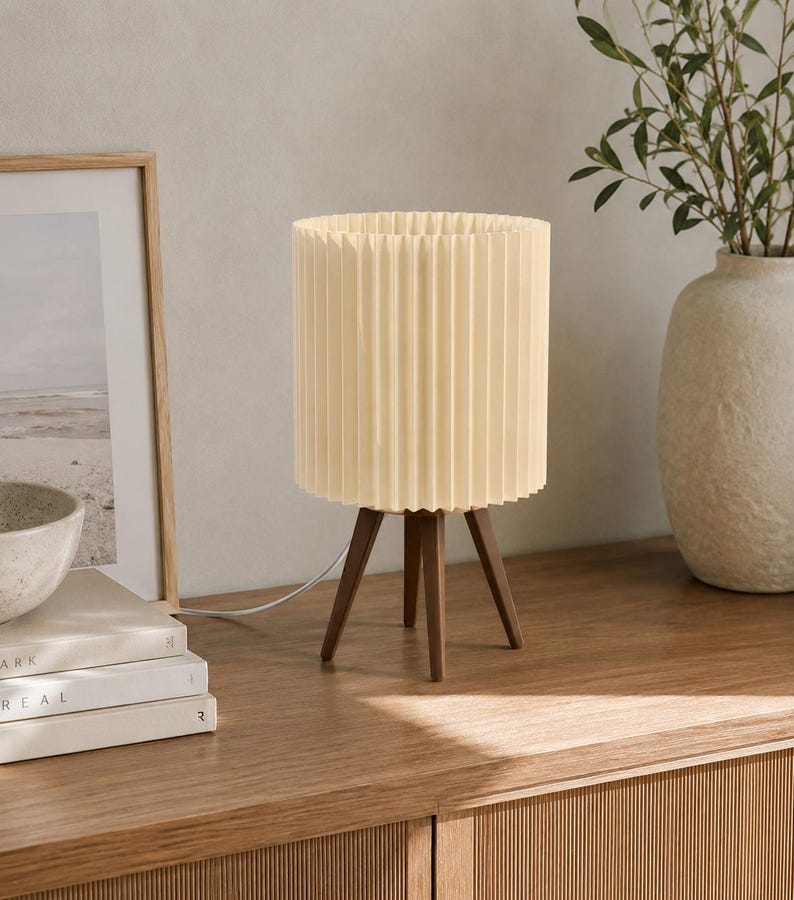 Puede incluir: Una l&aacute;mpara de mesa con una pantalla cil&iacute;ndrica plisada de color crema, sostenida por tres patas de madera de color marr&oacute;n oscuro. La l&aacute;mpara incluye un cable de alimentaci&oacute;n blanco que sale de la base, con un dise&ntilde;o estructurado.