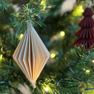May include: A close-up of a Christmas tree with a beige paper diamond-shaped ornament. The ornament is made of folded paper and is hanging from a green branch. A small, burgundy tree-shaped ornament is also visible.