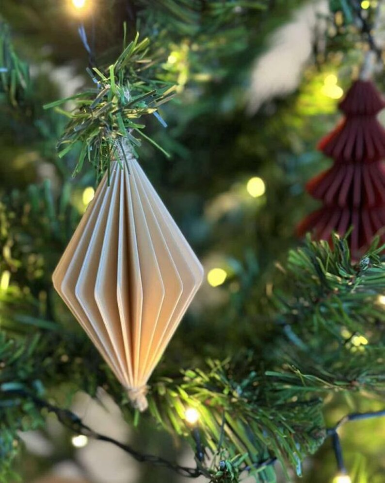 May include: A close-up of a Christmas tree with a beige paper diamond-shaped ornament. The ornament is made of folded paper and is hanging from a green branch. A small, burgundy tree-shaped ornament is also visible.