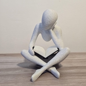 May include: A white, textured sculpture of a person seated cross-legged, reading a book. The figure's head rests on their hand, suggesting contemplation. The book is open, with white pages. The sculpture sits on a wooden surface.