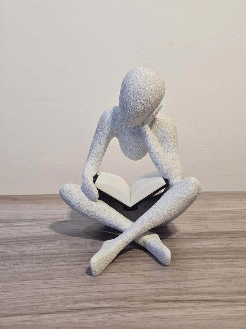 May include: A white, textured sculpture of a person seated cross-legged, reading a book. The figure's head rests on their hand, suggesting contemplation. The book is open, with white pages. The sculpture sits on a wooden surface.