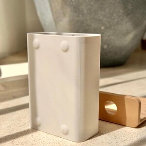 May include: A rectangular white plastic container with rounded corners and four small circular indentations on the front surface. Beside it sits a small, light tan L-shaped bracket featuring a single circular hole on its vertical side.