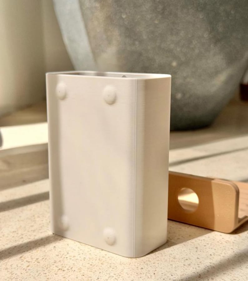 May include: A rectangular white plastic container with rounded corners and four small circular indentations on the front surface. Beside it sits a small, light tan L-shaped bracket featuring a single circular hole on its vertical side.
