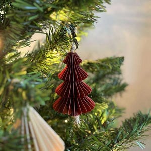 May include: A burgundy paper Christmas tree ornament hanging on a green artificial tree. The ornament is made of layered, folded paper, and has a string for hanging. The tree is decorated with other ornaments and lights.