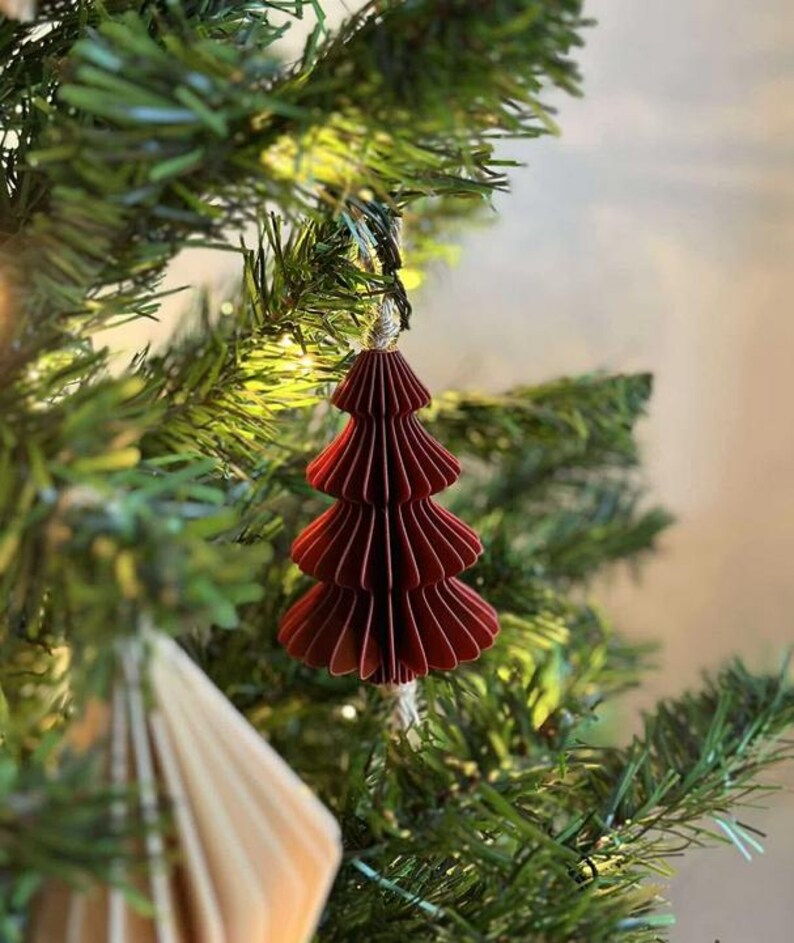 May include: A burgundy paper Christmas tree ornament hanging on a green artificial tree. The ornament is made of layered, folded paper, and has a string for hanging. The tree is decorated with other ornaments and lights.