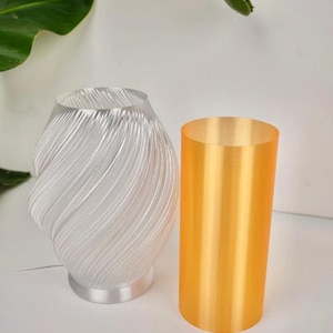 May include: A clear, swirled glass lamp with a silver base and a cylindrical orange lamp. The lamp has a unique, artistic design. The orange lamp is a solid, opaque cylinder. The background is white with a green leaf.