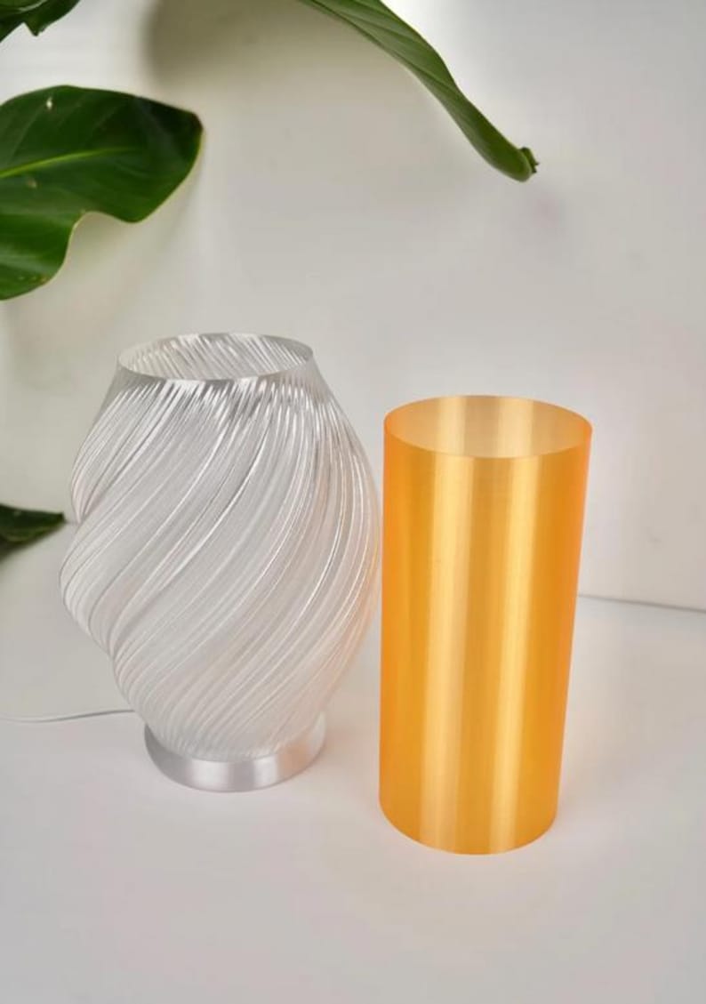 May include: A clear, swirled glass lamp with a silver base and a cylindrical orange lamp. The lamp has a unique, artistic design. The orange lamp is a solid, opaque cylinder. The background is white with a green leaf.