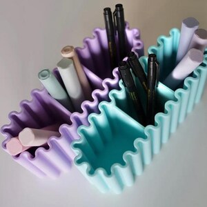 K&ouml;nnte beinhalten: Zwei wellenf&ouml;rmige Schreibtisch-Organizer in Pastellviolett und T&uuml;rkis, gef&uuml;llt mit Stiften und Bleistiften. Die Beh&auml;lter haben ein geriffeltes Design und sind in drei Abschnitte unterteilt. Die Stifte sind schwarz und die Bleistifte in verschiedenen Pastellfarben.