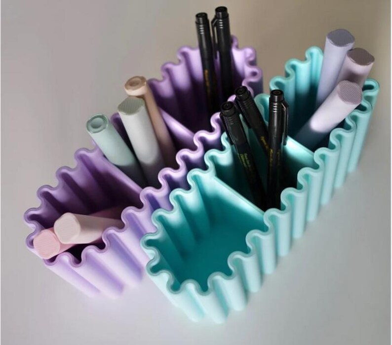 K&ouml;nnte beinhalten: Zwei wellenf&ouml;rmige Schreibtisch-Organizer in Pastellviolett und T&uuml;rkis, gef&uuml;llt mit Stiften und Bleistiften. Die Beh&auml;lter haben ein geriffeltes Design und sind in drei Abschnitte unterteilt. Die Stifte sind schwarz und die Bleistifte in verschiedenen Pastellfarben.