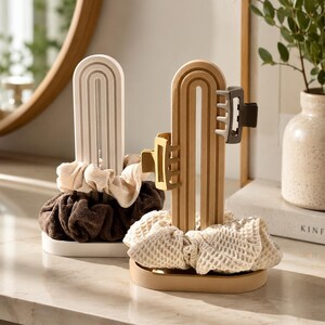May include: Two arched hair accessory organizers with oval bases. One is white, holding a cream ribbed scrunchie and a dark brown scrunchie. The other is tan, holding a beige waffle-textured scrunchie, a yellow hair claw, and a dark brown hair claw.