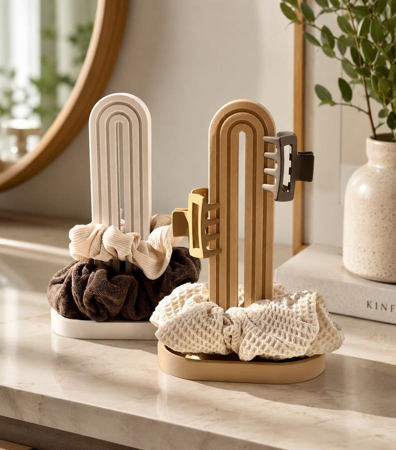 May include: Two arched hair accessory organizers with oval bases. One is white, holding a cream ribbed scrunchie and a dark brown scrunchie. The other is tan, holding a beige waffle-textured scrunchie, a yellow hair claw, and a dark brown hair claw.