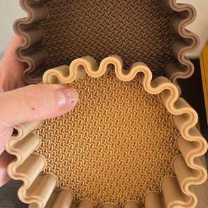 Könnte beinhalten: Zwei braune, wellenförmige, kreisförmige Tabletts. Die Innenseite jedes Tabletts hat eine strukturierte, gemusterte Oberfläche. Ein Tablett wird hochgehalten, wodurch die Unterseite des Tabletts sichtbar wird. Die Tabletts scheinen aus einem starren Material zu bestehen.