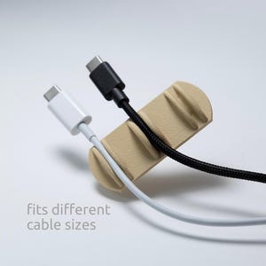 May include: A beige cable organiser with three distinct slots holding a white USB-C cable and a black braided USB-C cable. The organiser features a textured surface and the printed text "fits different cable sizes" in a light grey font.