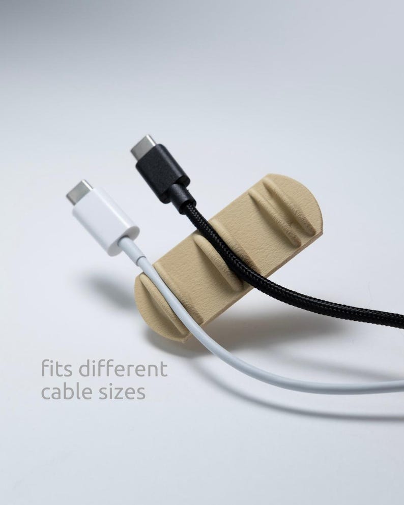 May include: A beige cable organiser with three distinct slots holding a white USB-C cable and a black braided USB-C cable. The organiser features a textured surface and the printed text "fits different cable sizes" in a light grey font.