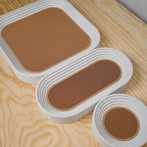 May include: A set of three decorative trays in varying sizes. The trays have a white, textured exterior with a brown, perforated interior. The shapes include a square, an oval, and a circle. The trays are arranged on a light wood surface.