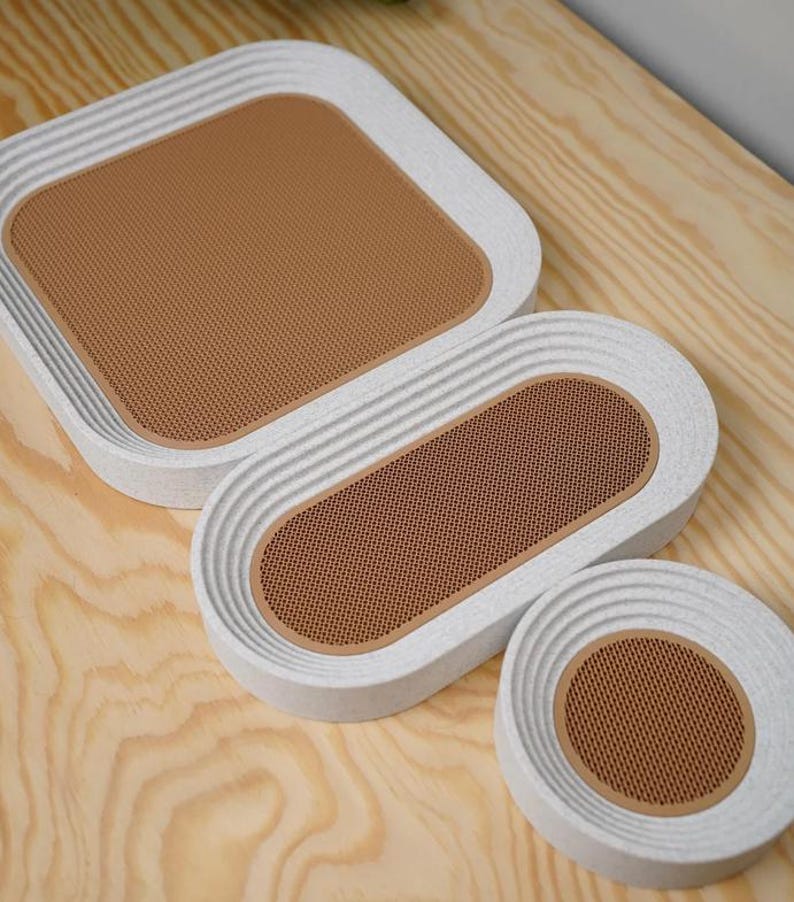 May include: A set of three decorative trays in varying sizes. The trays have a white, textured exterior with a brown, perforated interior. The shapes include a square, an oval, and a circle. The trays are arranged on a light wood surface.