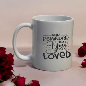 May include: White ceramic mug with a large handle. The mug features the text "little REMINDER that You are LOVED" in black script with red heart accents. The mug is surrounded by red roses.