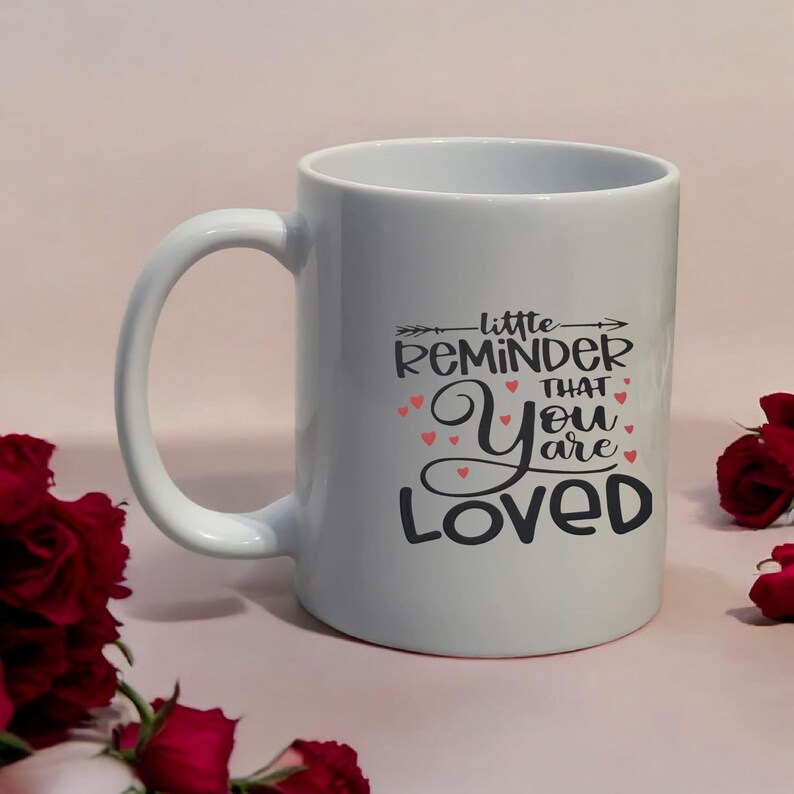 May include: White ceramic mug with a large handle. The mug features the text "little REMINDER that You are LOVED" in black script with red heart accents. The mug is surrounded by red roses.