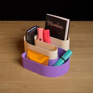 May include: A desk organiser with a wavy, textured design featuring a cream-coloured upper section and a purple lower section. It holds a dark notebook with the text "Microalmas", two pink highlighters, a dark wallet, a gold envelope, and a teal pen.