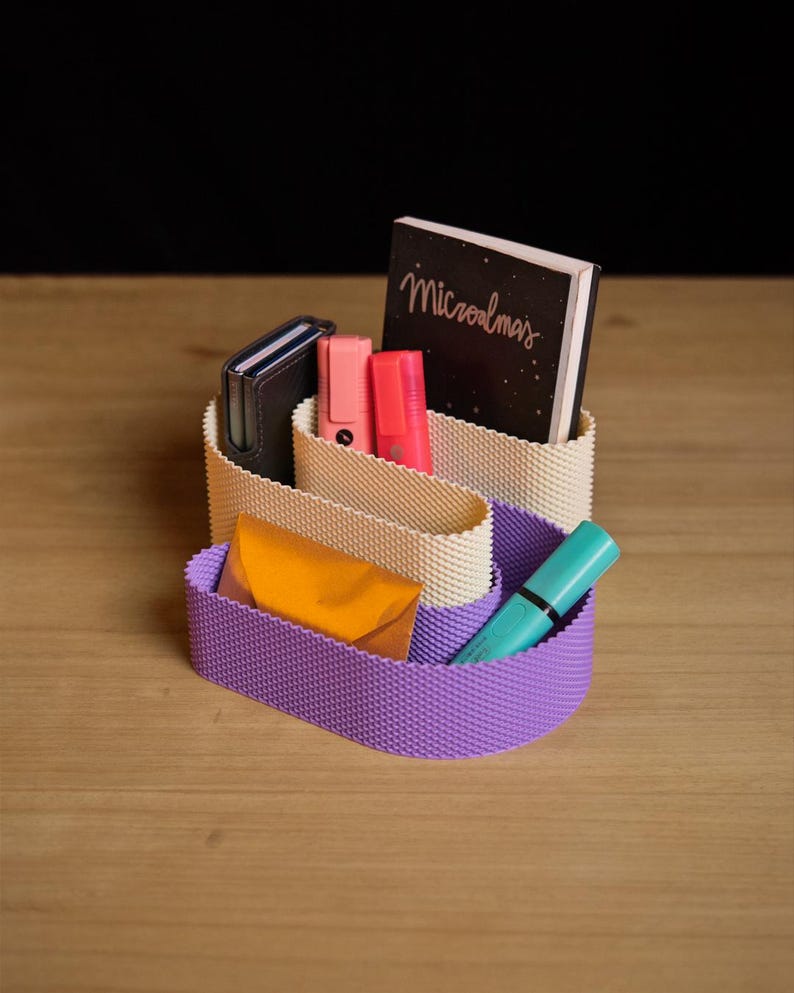 May include: A desk organiser with a wavy, textured design featuring a cream-coloured upper section and a purple lower section. It holds a dark notebook with the text "Microalmas", two pink highlighters, a dark wallet, a gold envelope, and a teal pen.