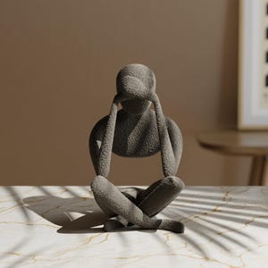 K&ouml;nnte beinhalten: Eine graue Skulptur einer Person in sitzender, meditativer Pose, mit den H&auml;nden &uuml;ber dem Kopf. Die strukturierte Oberfl&auml;che der Figur kontrastiert mit der glatten, gemusterten Oberfl&auml;che, auf der sie ruht. Die Skulptur ist ein dekorativer Gegenstand.