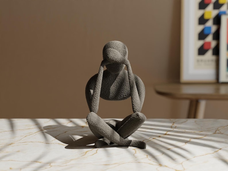 K&ouml;nnte beinhalten: Eine graue Skulptur einer Person in sitzender, meditativer Pose, mit den H&auml;nden &uuml;ber dem Kopf. Die strukturierte Oberfl&auml;che der Figur kontrastiert mit der glatten, gemusterten Oberfl&auml;che, auf der sie ruht. Die Skulptur ist ein dekorativer Gegenstand.