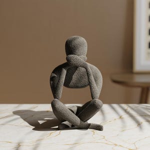 K&ouml;nnte beinhalten: Eine graue Skulptur einer Person in sitzender, meditativer Pose. Die Figur hat die Arme verschr&auml;nkt, eine Hand ber&uuml;hrt das Kinn. Die Skulptur steht auf einer Marmoroberfl&auml;che mit neutralem Hintergrund.