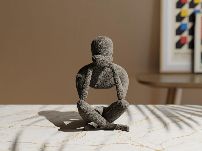 K&ouml;nnte beinhalten: Eine graue Skulptur einer Person in sitzender, meditativer Pose. Die Figur hat die Arme verschr&auml;nkt, eine Hand ber&uuml;hrt das Kinn. Die Skulptur steht auf einer Marmoroberfl&auml;che mit neutralem Hintergrund.