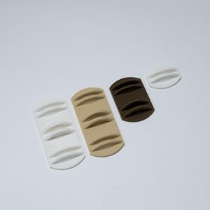May include: A collection of four small, rectangular and oval-shaped plastic organisers with raised, curved ridges. The set includes one long white piece, one long beige piece, one shorter dark brown piece, and one small white oval piece.