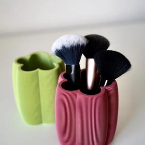 May include: A pink and a green pepper-shaped makeup brush holder with vertical lines. The pink holder contains several makeup brushes with black and white bristles. The holders are made of a ribbed material.