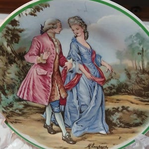 May include: A decorative plate with a painted scene of a couple in 18th-century clothing. The man is in a pink coat, the woman in a blue dress. The plate has a green border and a signature at the bottom, reminiscent of a period piece.