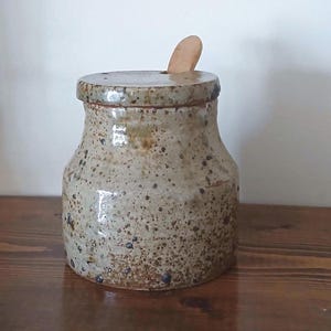 Puede incluir: Un tarro de cerámica moteada con tapa y una pequeña cuchara de madera. El tarro es de color beige claro con motas marrones oscuras y un acabado ligeramente brillante. La tapa tiene un pequeño agujero para la cuchara.