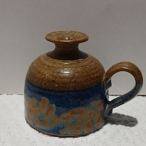 May include: Handmade ceramic mug with a unique design. The mug features a brown and blue colour scheme with a handle. The mug has a bulbous body and a small, rounded top. The mug is likely used for drinking beverages.