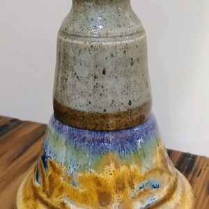 Peut inclure: Un vase en céramique fait à la main avec un design unique en couches. Le vase présente une base dans les tons de jaune et de marron, passant au bleu et au vert, et surmontée d'une section grise et marron. Le bord supérieur est marron.
