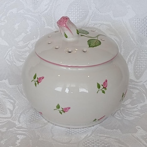 Peut inclure: Un pot en céramique blanc avec un couvercle, orné de roses roses et de feuilles vertes. Le couvercle comporte une poignée en forme de bouton de rose rose et de petits trous. Une ligne rose accentue le bord, créant un récipient charmant sur le thème floral.