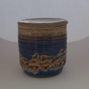 May include: A ceramic jar with a lid, cylindrical in shape. The jar has a blue base with brown and tan detailing. The upper section is tan, and the lid matches. The jar is likely handmade and suitable for storage of dry goods.