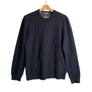 May include: A dark navy blue long-sleeve sweater featuring a vertical cable knit pattern on the front panel and a ribbed crew neckline. The interior collar displays a purple and teal geometric pattern, and the garment hangs on a wooden hanger.