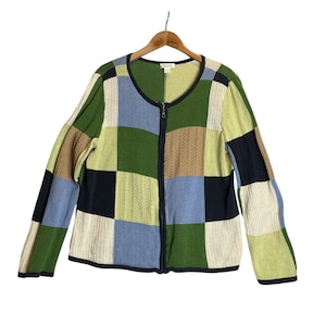 May include: A patchwork cardigan sweater with a zip-up front. The sweater features a variety of colors, including green, blue, beige, and black, arranged in a square pattern. The sweater is hanging on a wooden hanger.