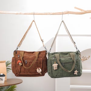 Puede incluir: Dos bolsos tote de pana, uno marrón y otro verde, colgados de una rama. Cada bolso tiene una correa estampada, múltiples bolsillos y adornos decorativos. El bolso marrón tiene un adorno de casa y pájaro, el verde un adorno de taza de café.