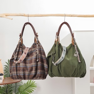 May include: Two hobo-style shoulder bags hanging from a branch. One bag features a brown plaid pattern, while the other is solid olive green with a corduroy texture. Both bags include brown leather-look handles and decorative patterned straps.