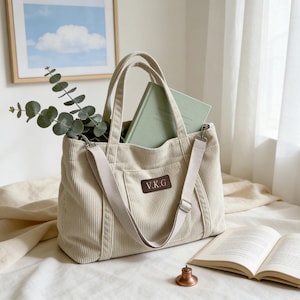 May include: A cream corduroy tote bag with a shoulder strap, containing a green book and eucalyptus sprigs. The bag features a rectangular patch with the initials "V.K.G." An open book and a small bell are on the bed.