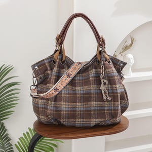 Può includere: Una borsa hobo a quadri marroni e blu con un'ampia tracolla decorativa con ricami floreali. La borsa presenta manici in similpelle marrone scuro, occhielli in metallo e un ciondolo pendente attaccato alla minuteria laterale.